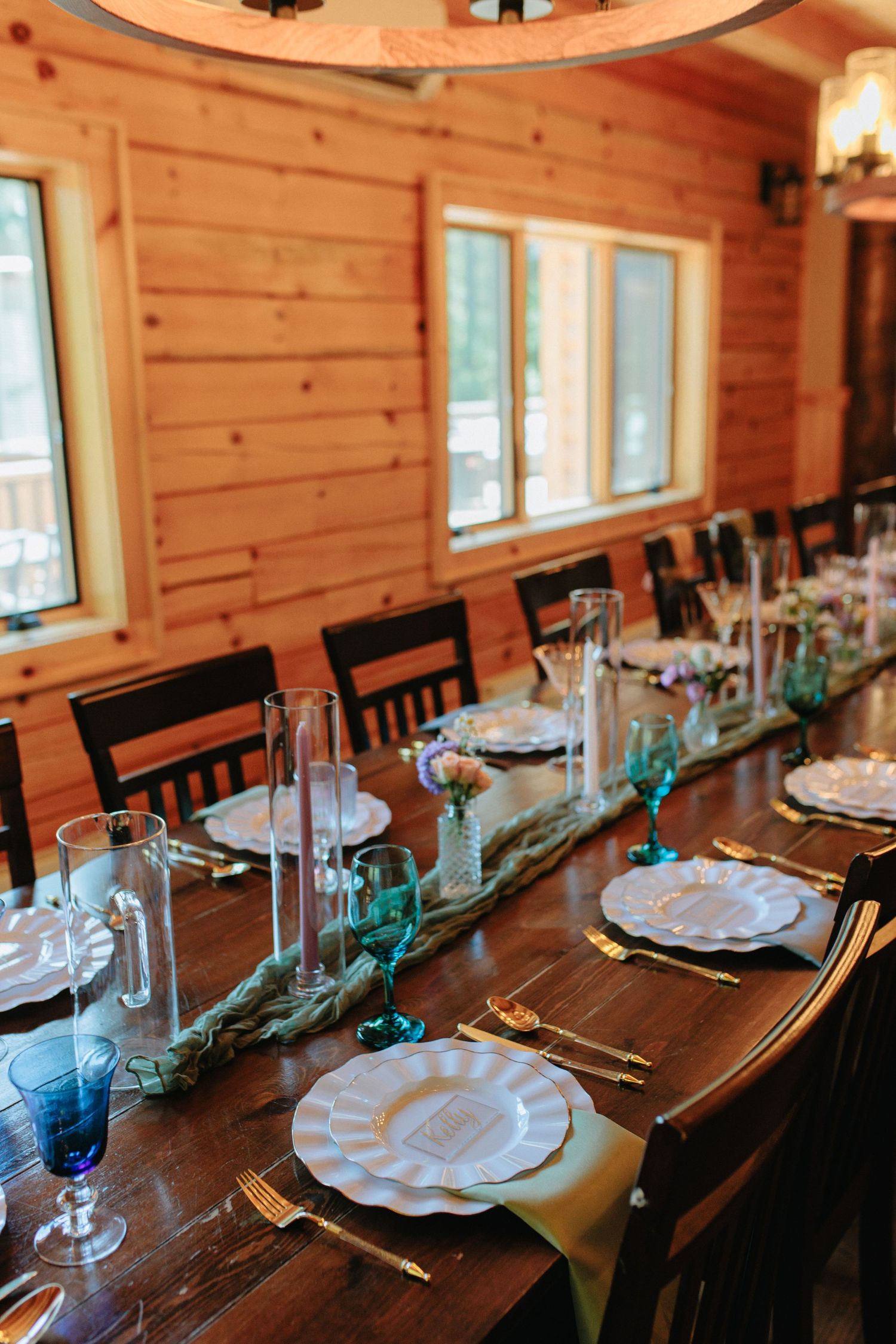 A formal table setting with white plates and blue glassware in a wooden cabin interior with natural lighting.