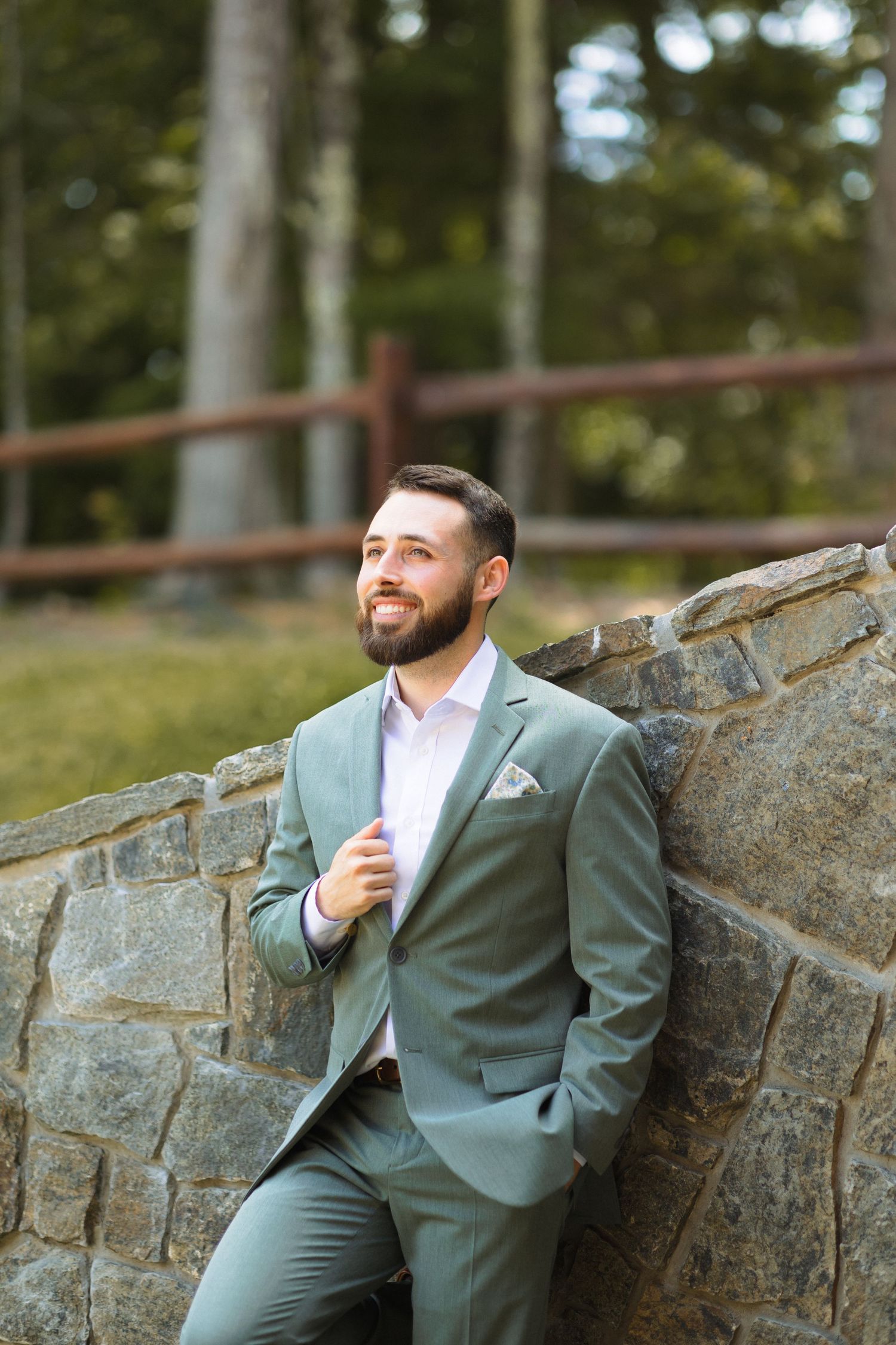 A wedding party member in a sage green suit leans against a rustic stone wall with a warm smile.