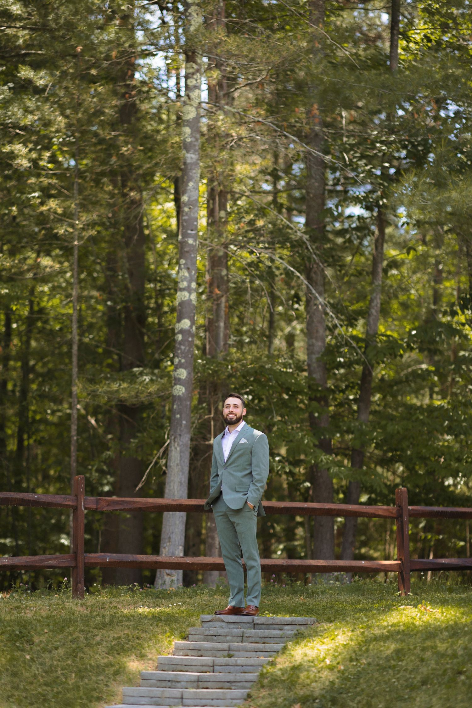 A person in a light colored suit stands on stone steps with a wooden fence and forest backdrop.