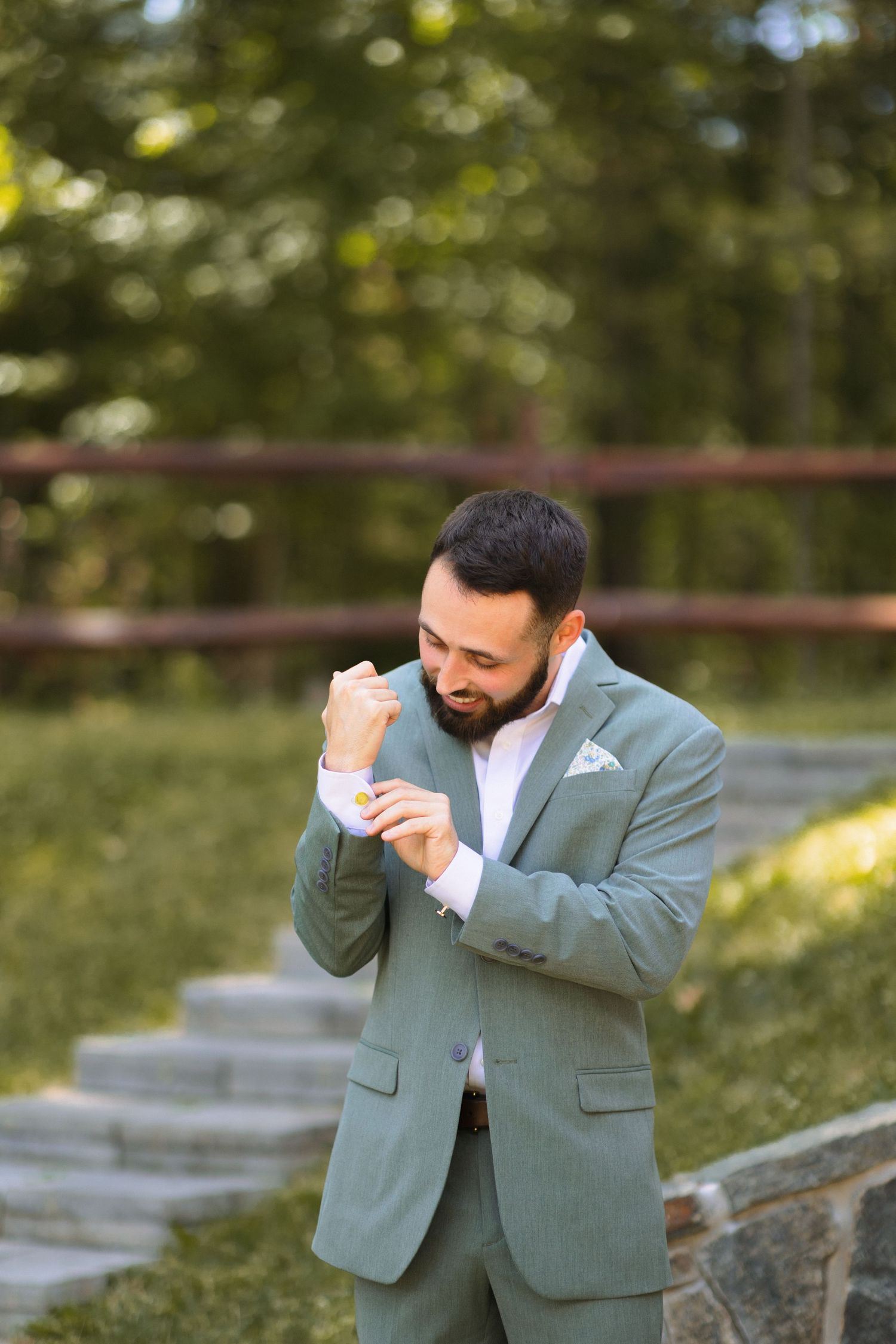 Person in a sage green suit adjusting their cufflinks outdoors near stone steps and greenery.