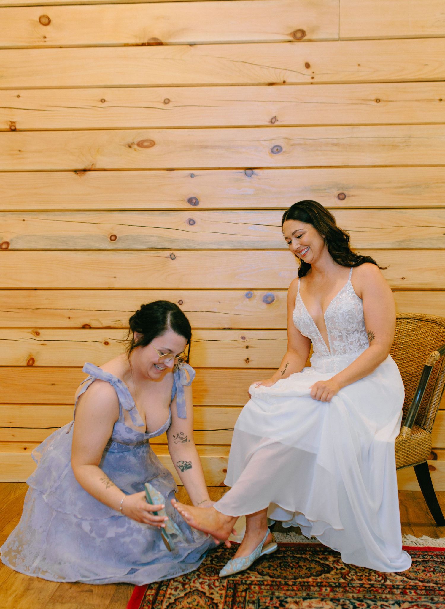 Two women share a tender moment as one helps the other with her shoes against a rustic wooden wall backdrop.