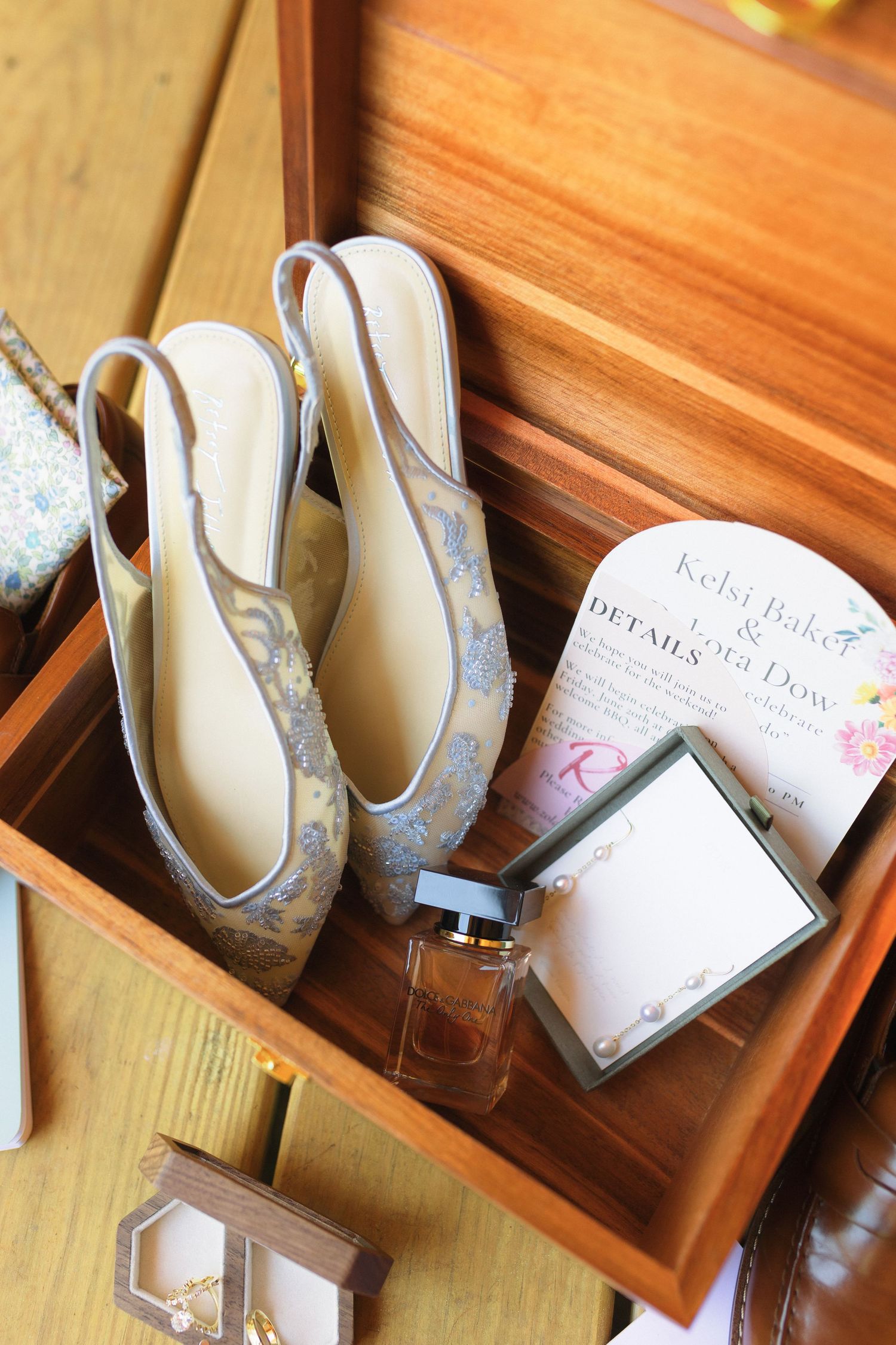Wedding accessories including silver shoes, perfume bottle and invitation details arranged in a wooden box.