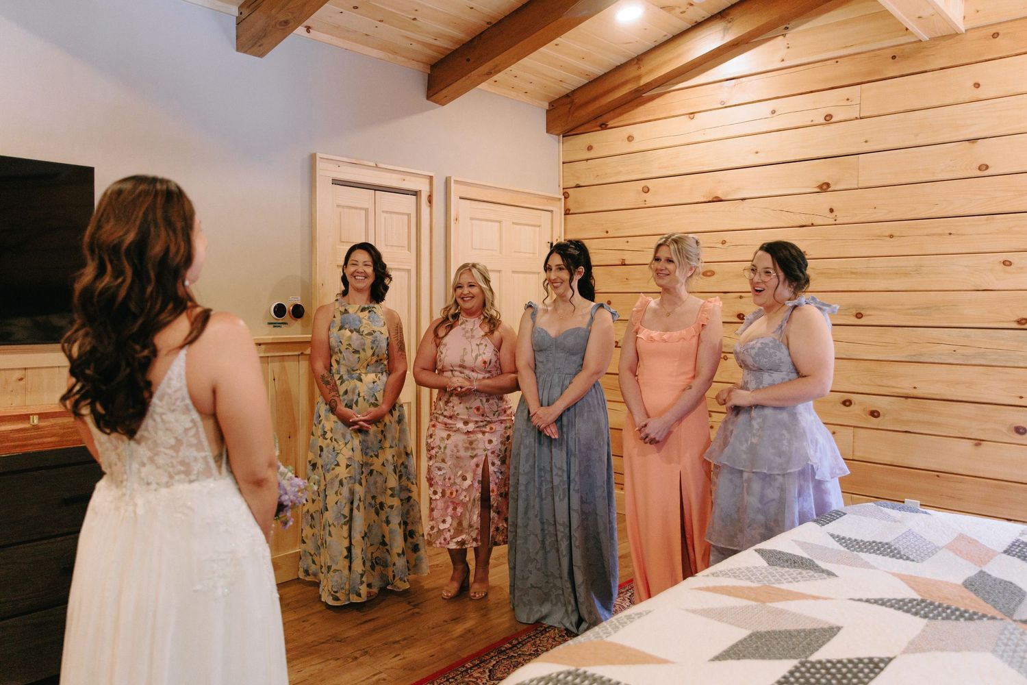 Bridesmaids in pastel floral dresses gather in a rustic wooden room with exposed beams.
