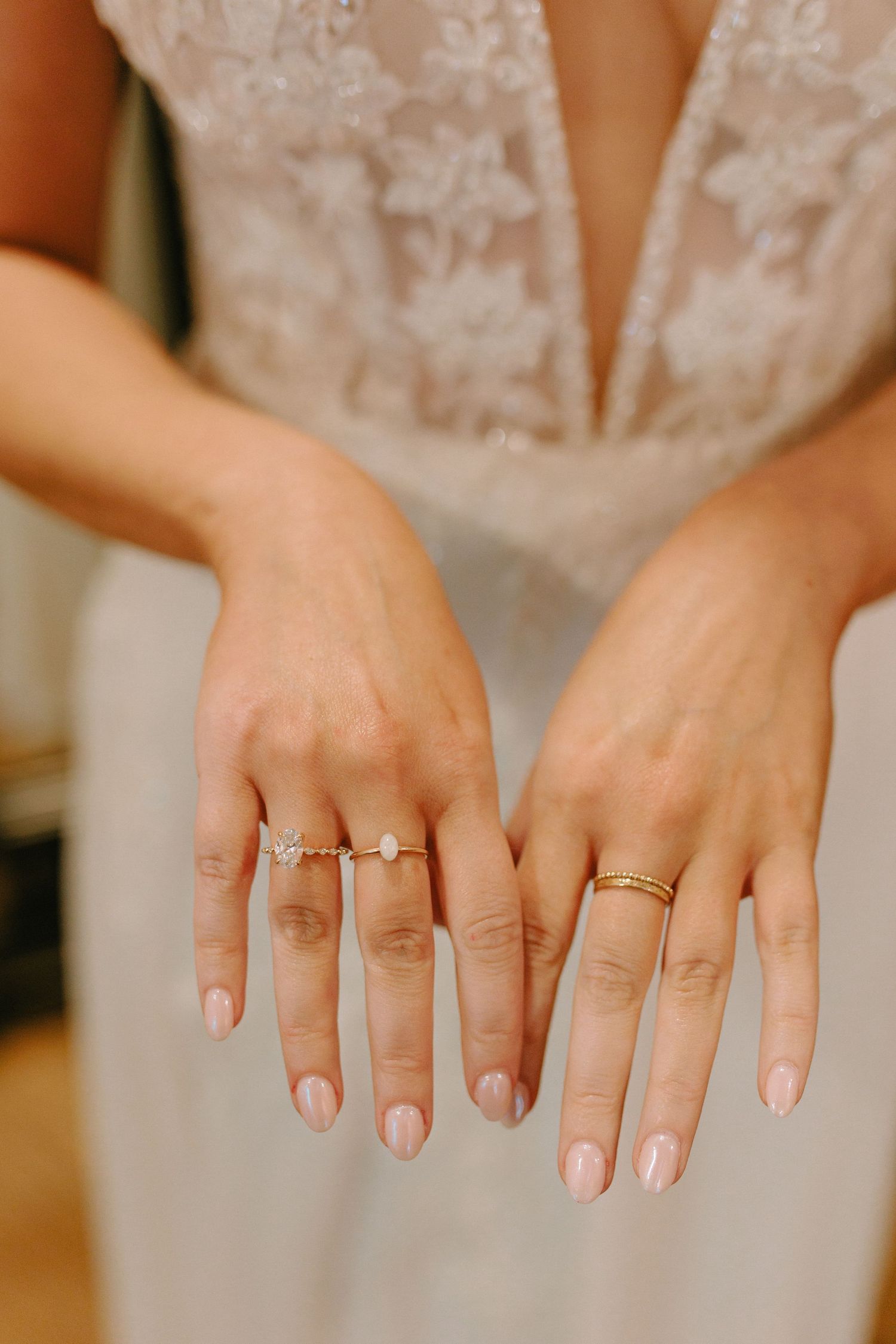 Close-up of hands wearing delicate wedding and engagement rings with natural manicure against white lace dress.
