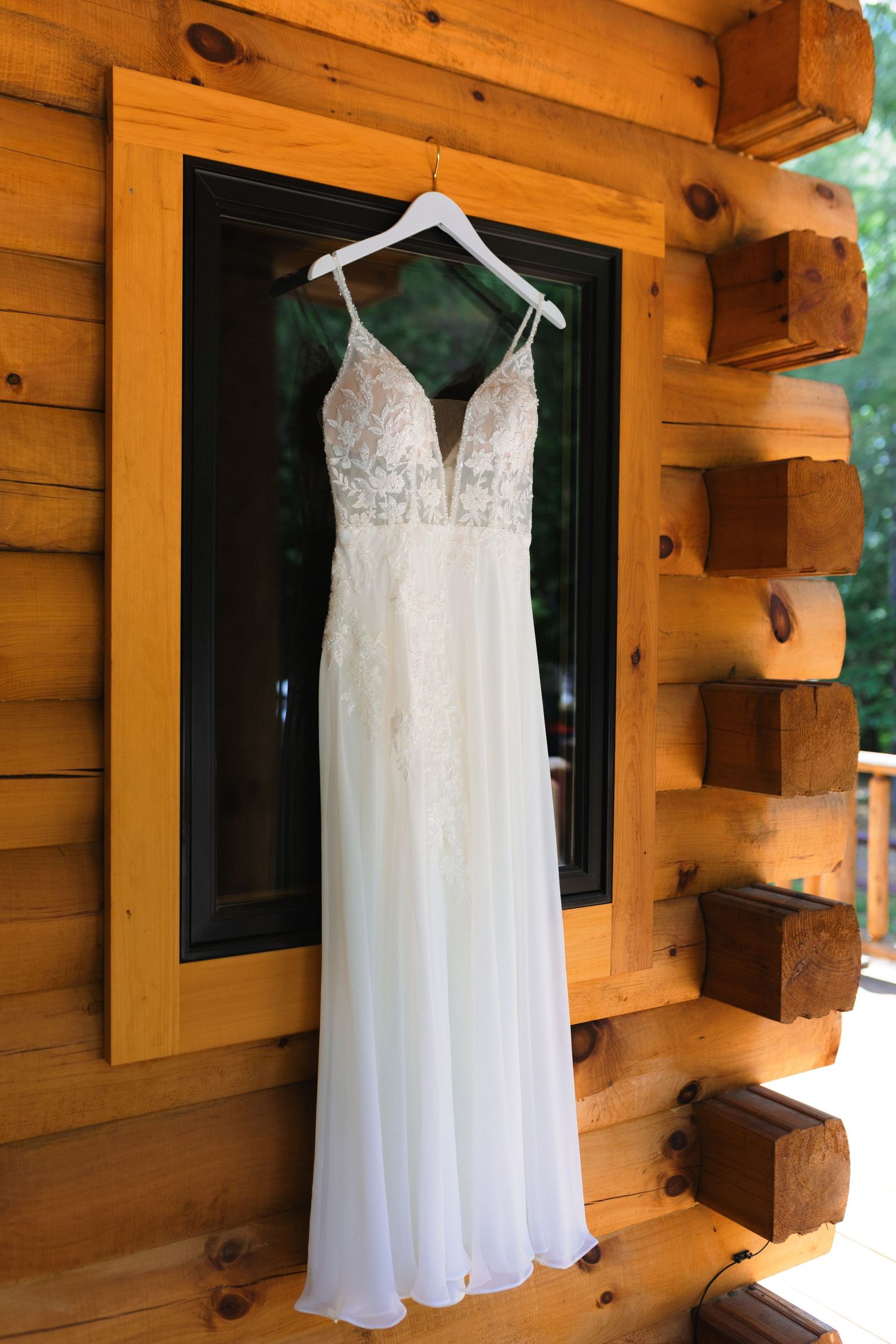Elegant white lace wedding dress with plunging neckline hanging on wooden cabin wall.