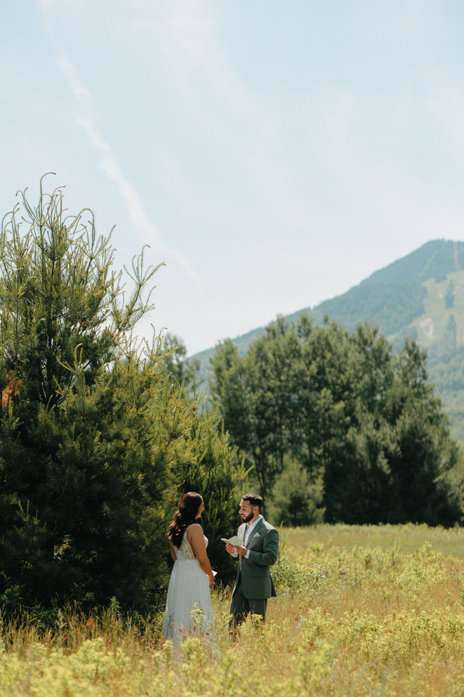 A couple shares an intimate moment in a mountain meadow with tall grass and evergreen trees in the background.