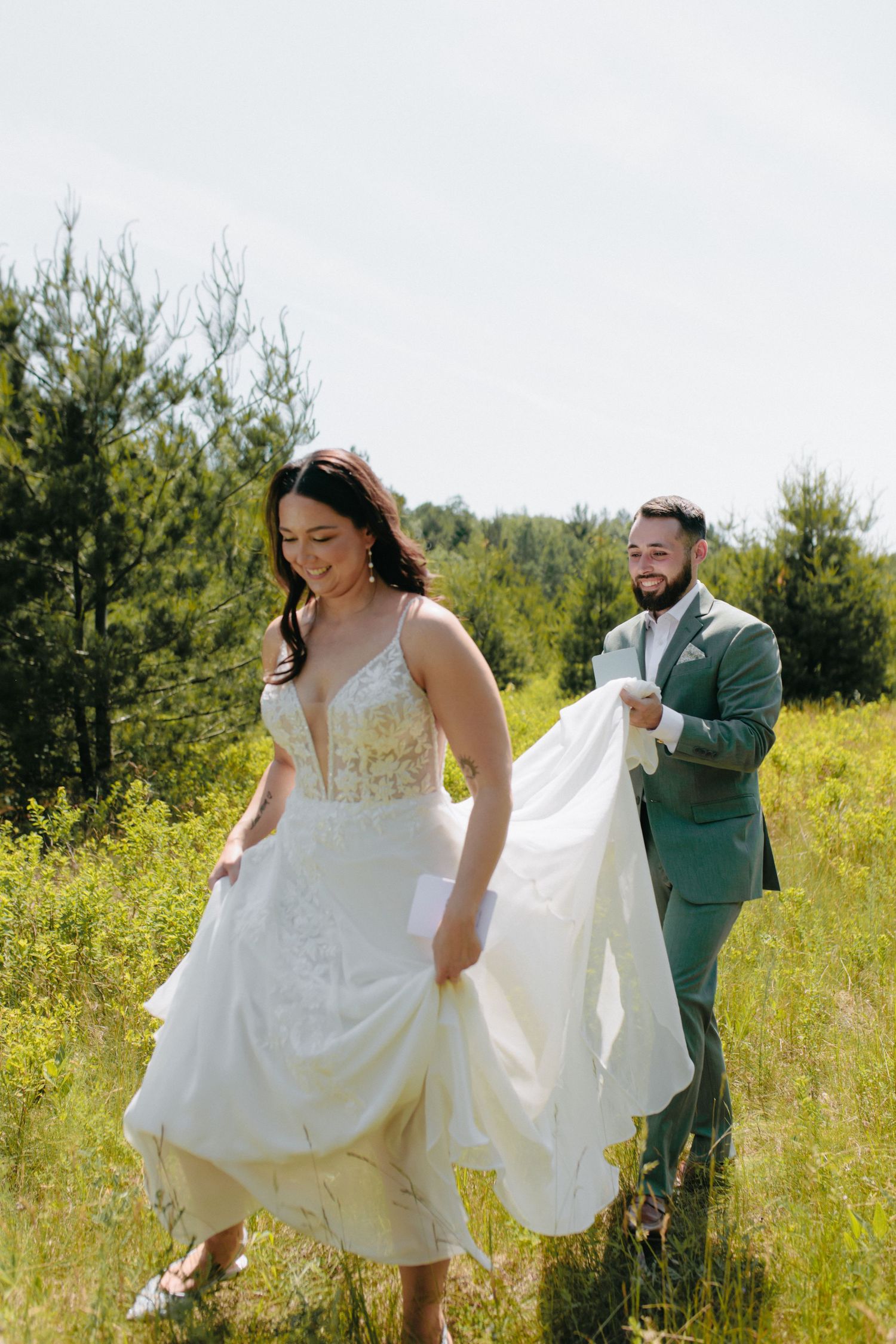 A couple walks through a sunny meadow with the bride holding her flowing white wedding dress in a playful moment.