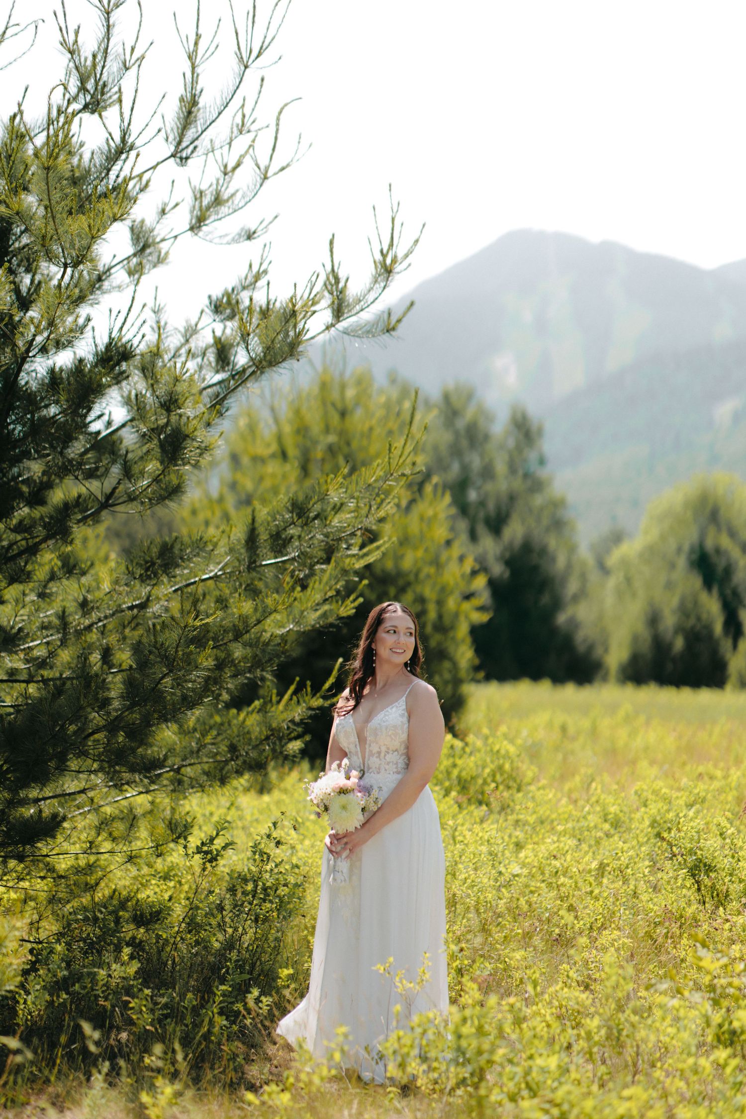 A bride poses in a flowing white dress among yellow wildflowers with mountains in the background.