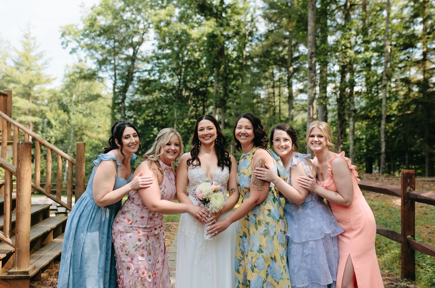 Group wearing pastel dresses raises champagne glasses in a toast during outdoor celebration in wooded setting.