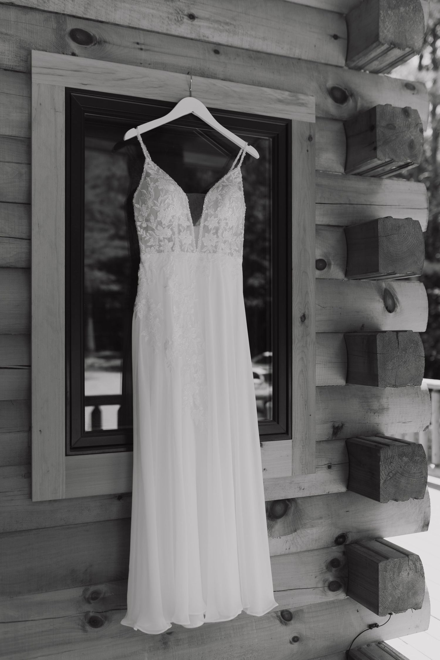 An elegant white wedding dress with lace bodice and plunging neckline hangs on display against a wooden cabin wall.