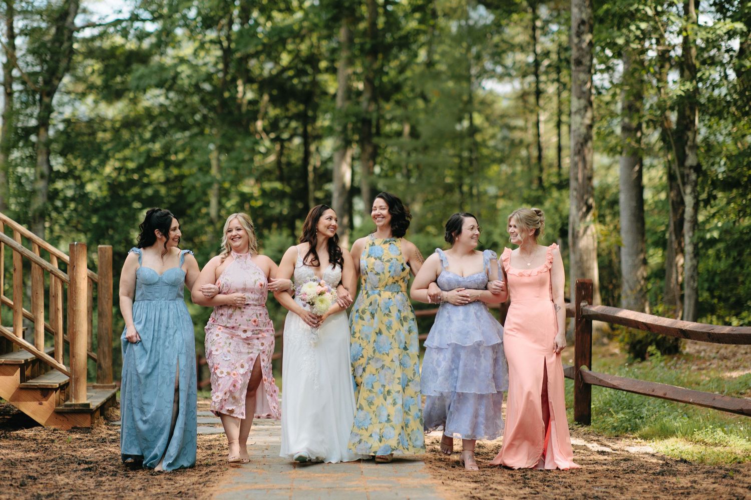 A wedding party in pastel dresses walks together on a wooded path.