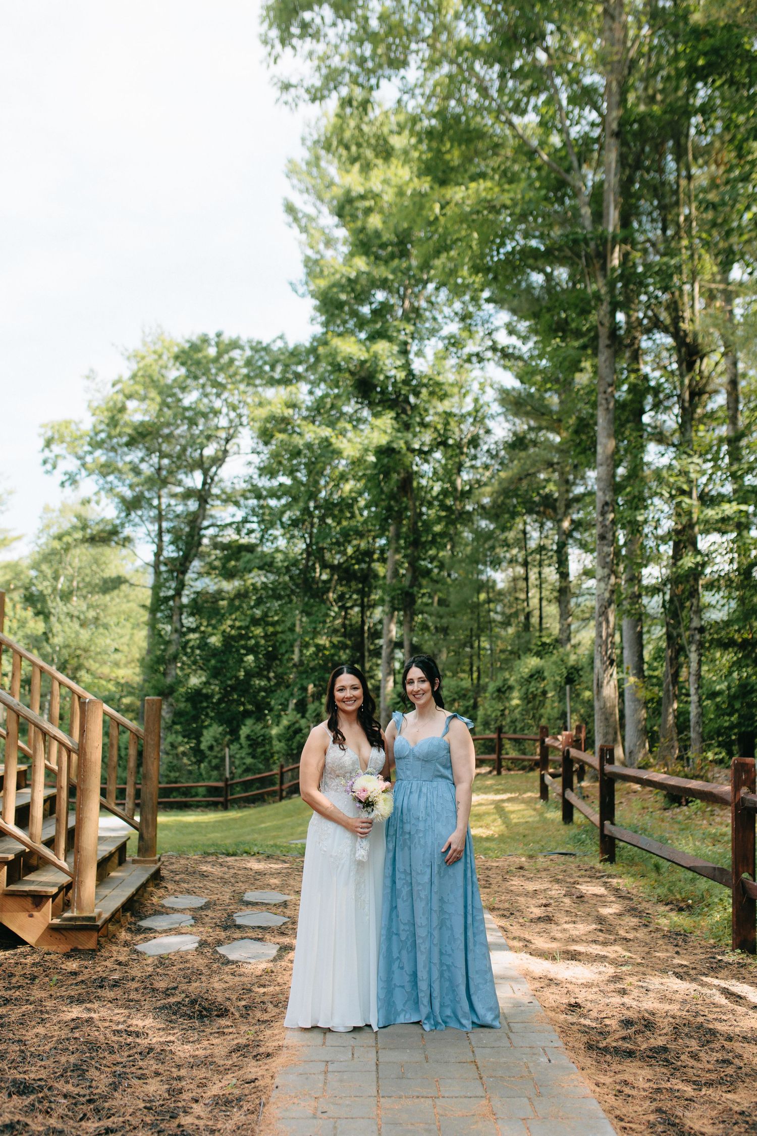 Two women in formal dresses stand together on a wooden walkway surrounded by a forest setting.
