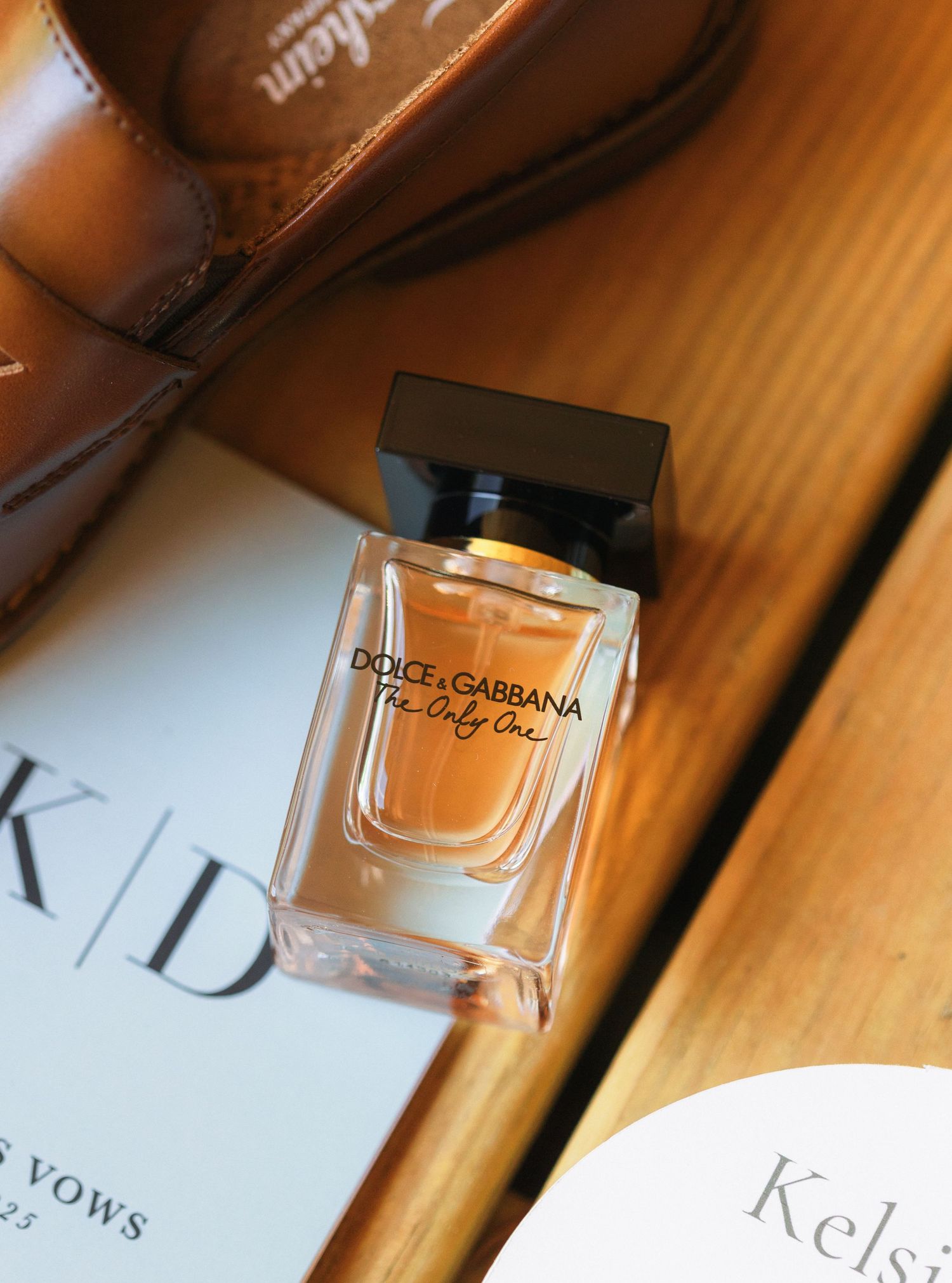 Dolce & Gabbana perfume bottle next to brown leather shoes and wedding stationery.