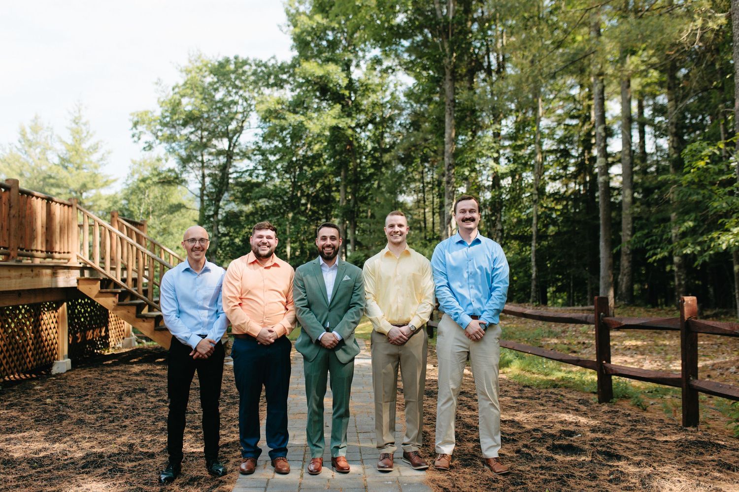 A group of people wearing pastel colored shirts stand together outdoors in front of a wooded area.
