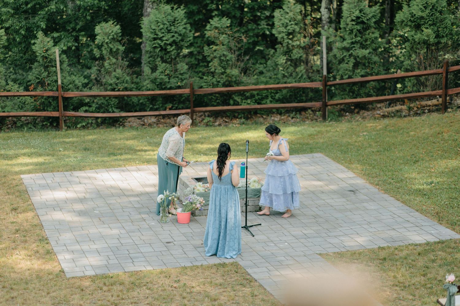 Three people in blue dresses performing a ceremony on a stone patio surrounded by a rustic wooden fence and greenery.