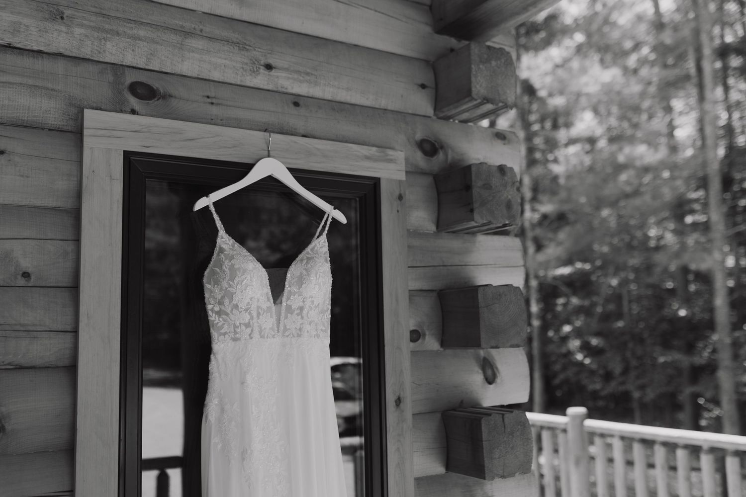 A white lace wedding dress hangs on display on a rustic wooden porch with trees visible in the background.