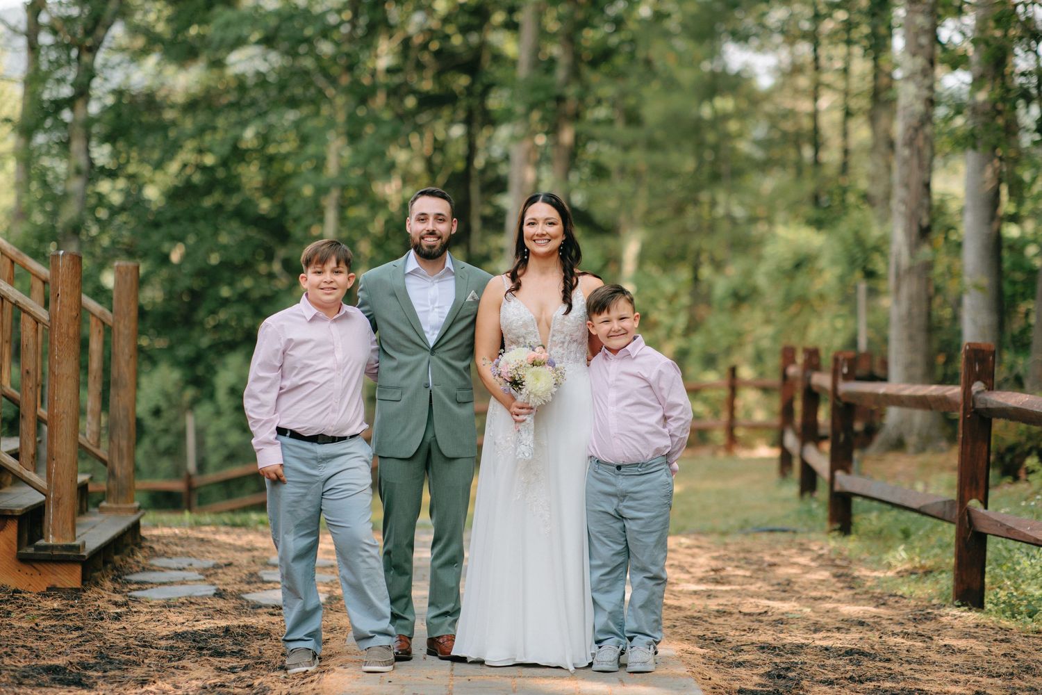 Family group in wedding attire poses together on a rustic woodland path with wooden fencing and green trees.
