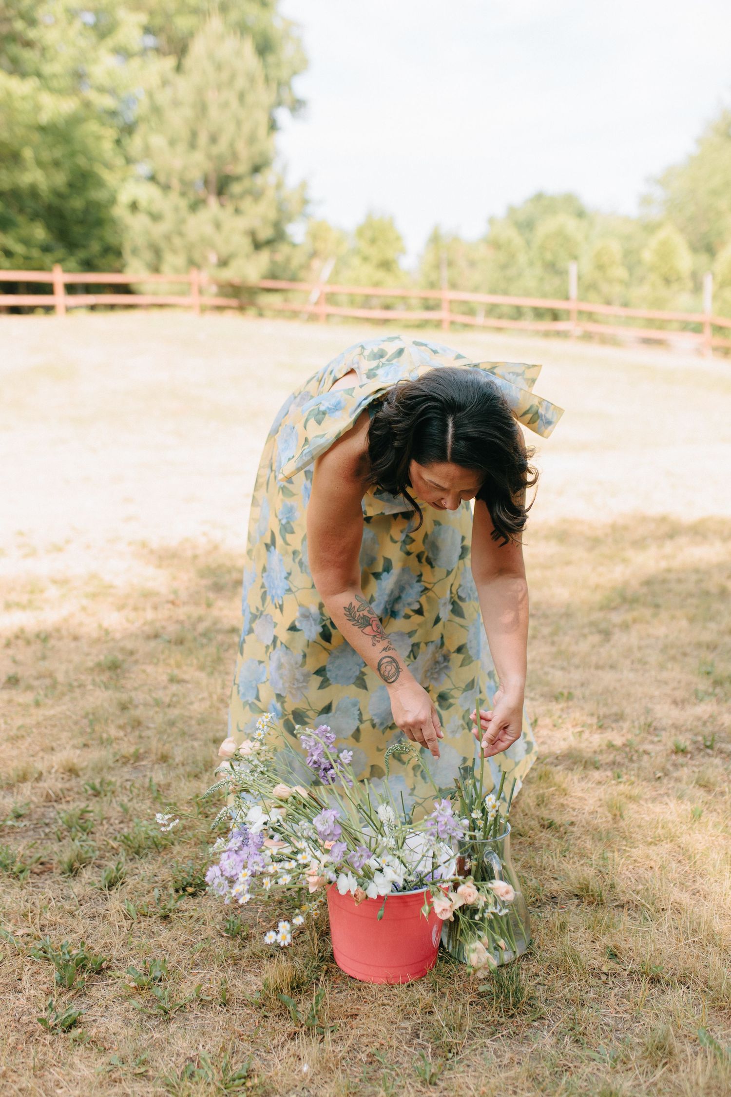 Person in a floral dress bends down to pick wildflowers in a sunny meadow.