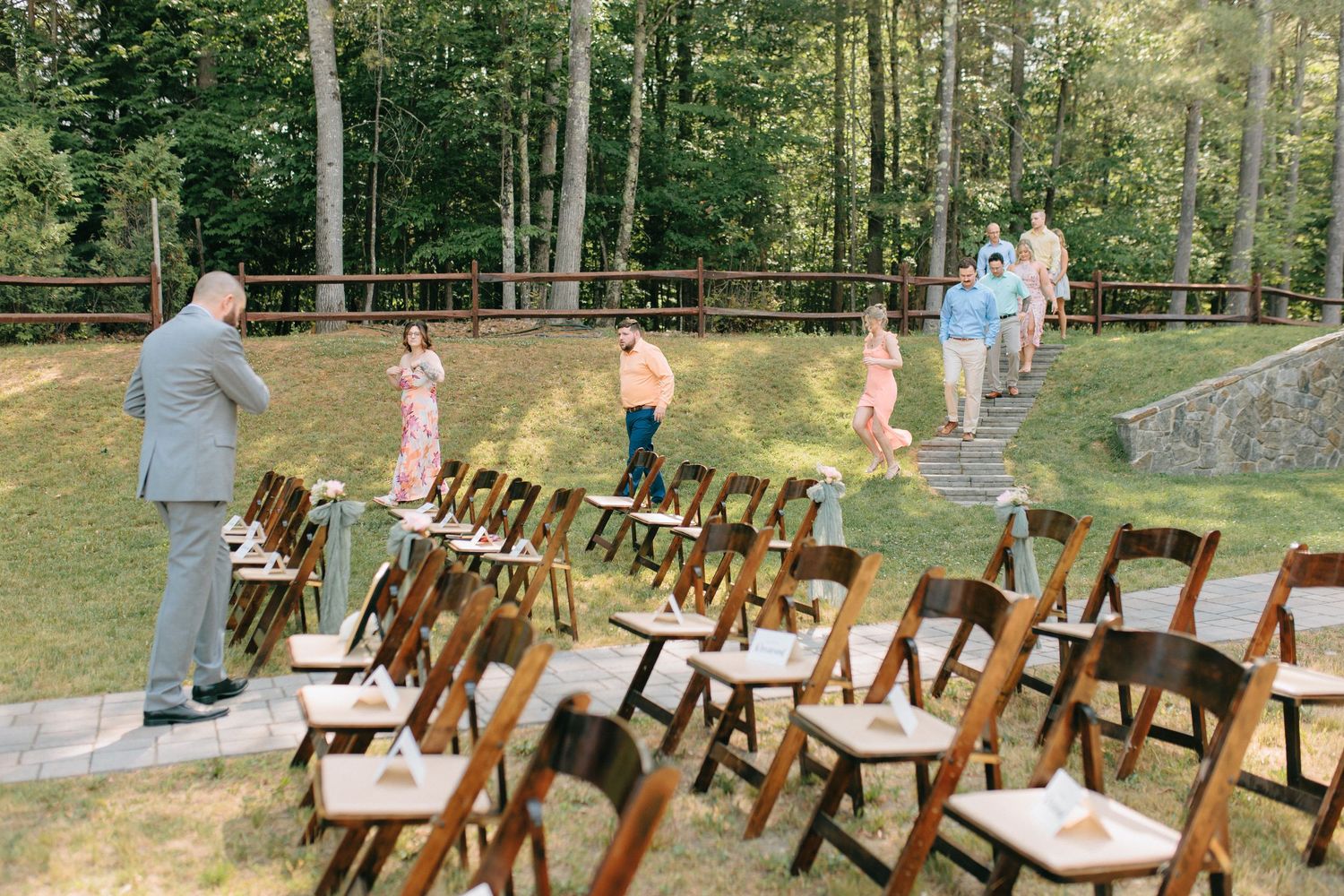 Wooden folding chairs arranged outdoors for a ceremony with people walking on a grassy hill in a wooded setting.