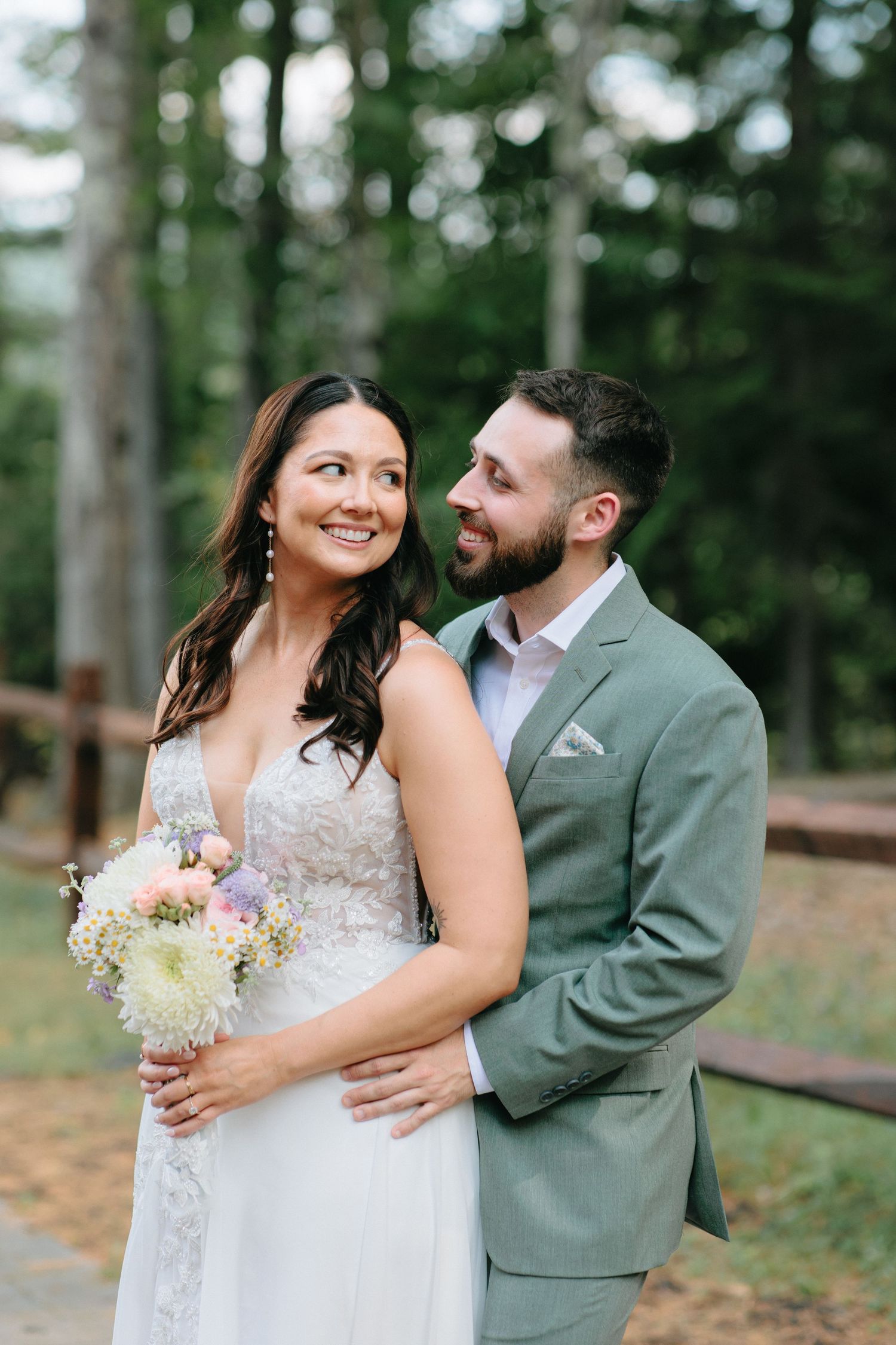 Newlyweds share an intimate moment in a forest setting with the bride holding a delicate floral bouquet.