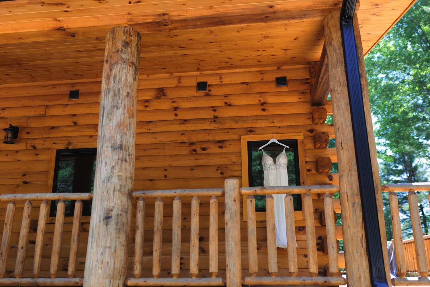A white wedding dress hangs on display outside a rustic log cabin porch with wooden railings and natural timber posts.