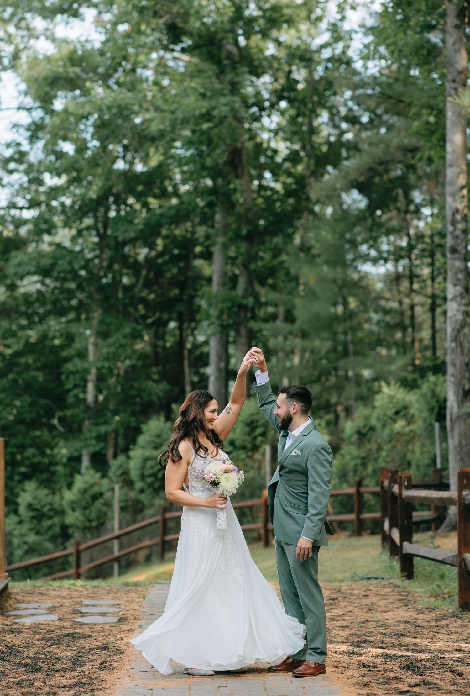 Couple dancing together on a woodland path, bride in white gown and groom in sage green suit surrounded by trees.