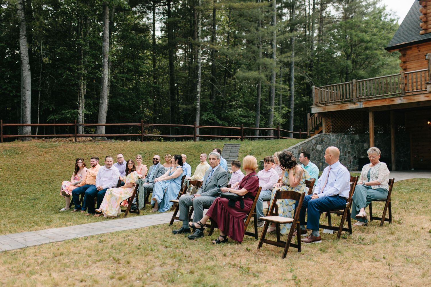 Large group sitting together on wooden benches outdoors with a rustic cabin and pine forest in background.
