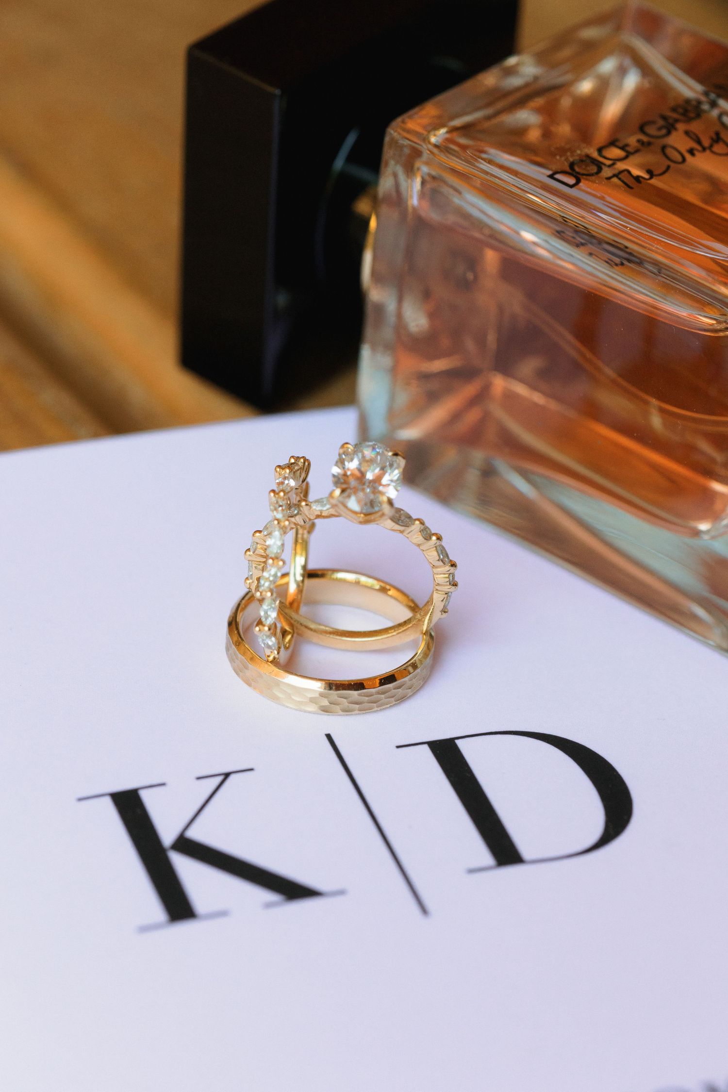 Gold wedding rings displayed on white paper with K|D initials next to perfume bottle.