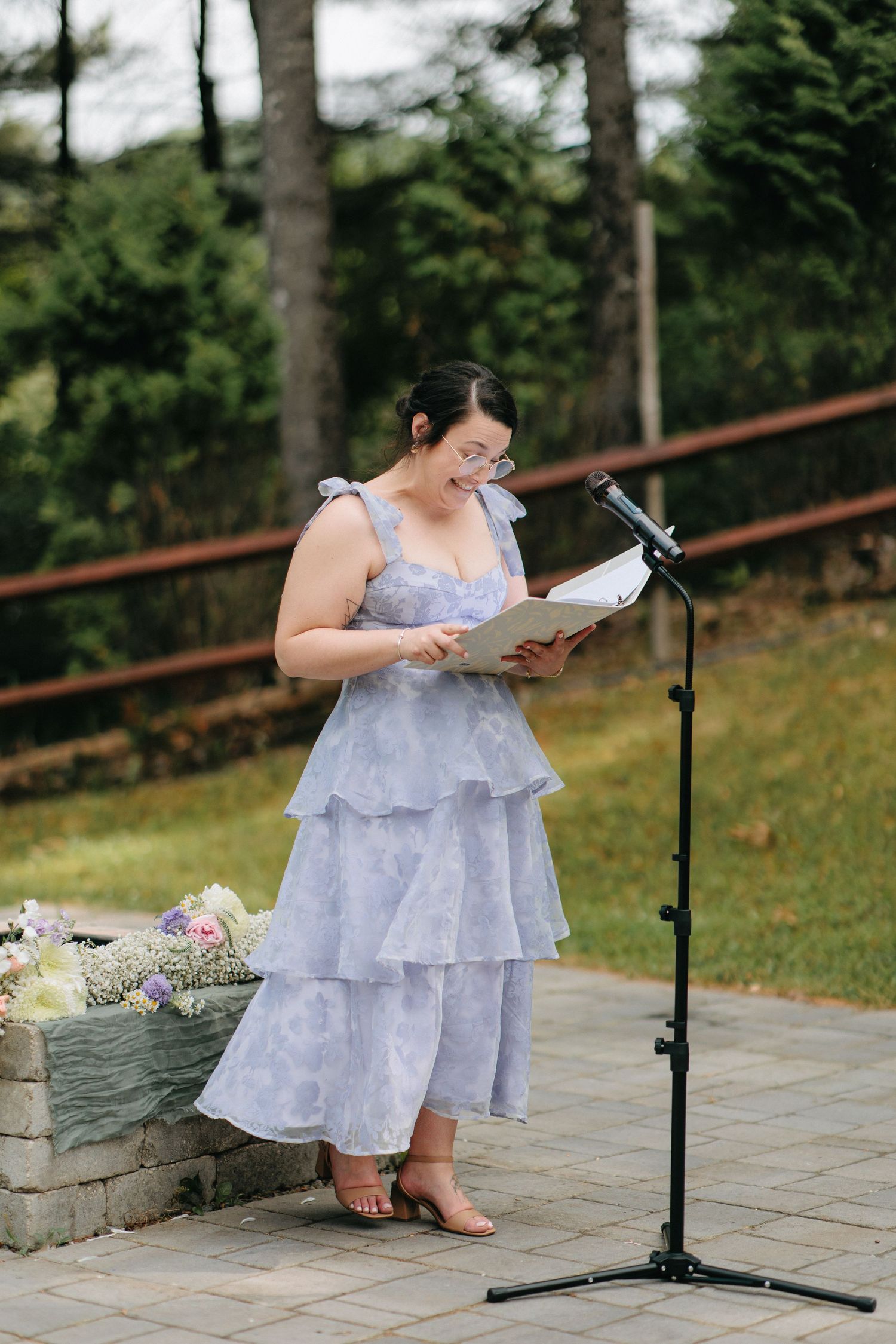 Someone in a light blue flowy dress reads from papers while sitting outdoors by a stone wall on a paved terrace.