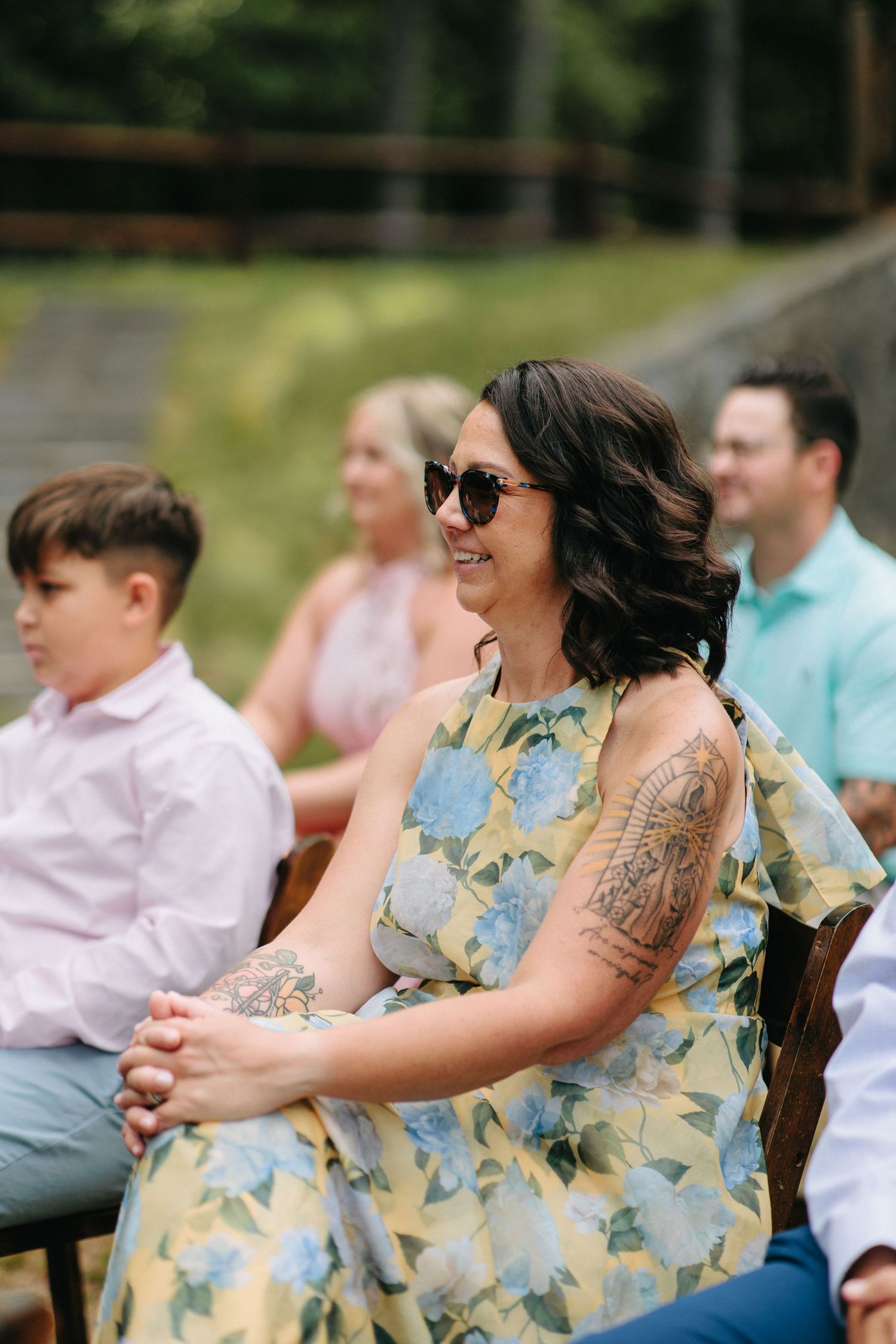 Wedding guest in a floral dress with sunglasses sits outdoors during a summer ceremony.