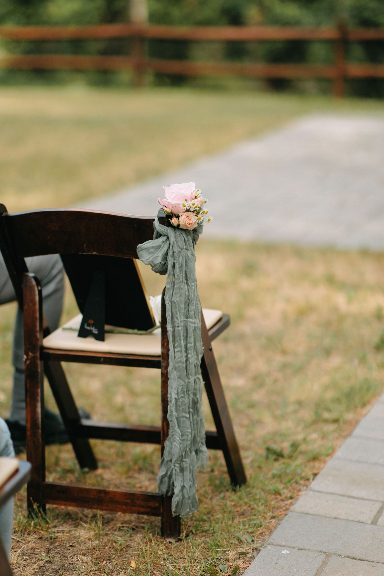 Rustic wooden ceremony chairs decorated with sage green fabric and pink floral accents line a garden path.