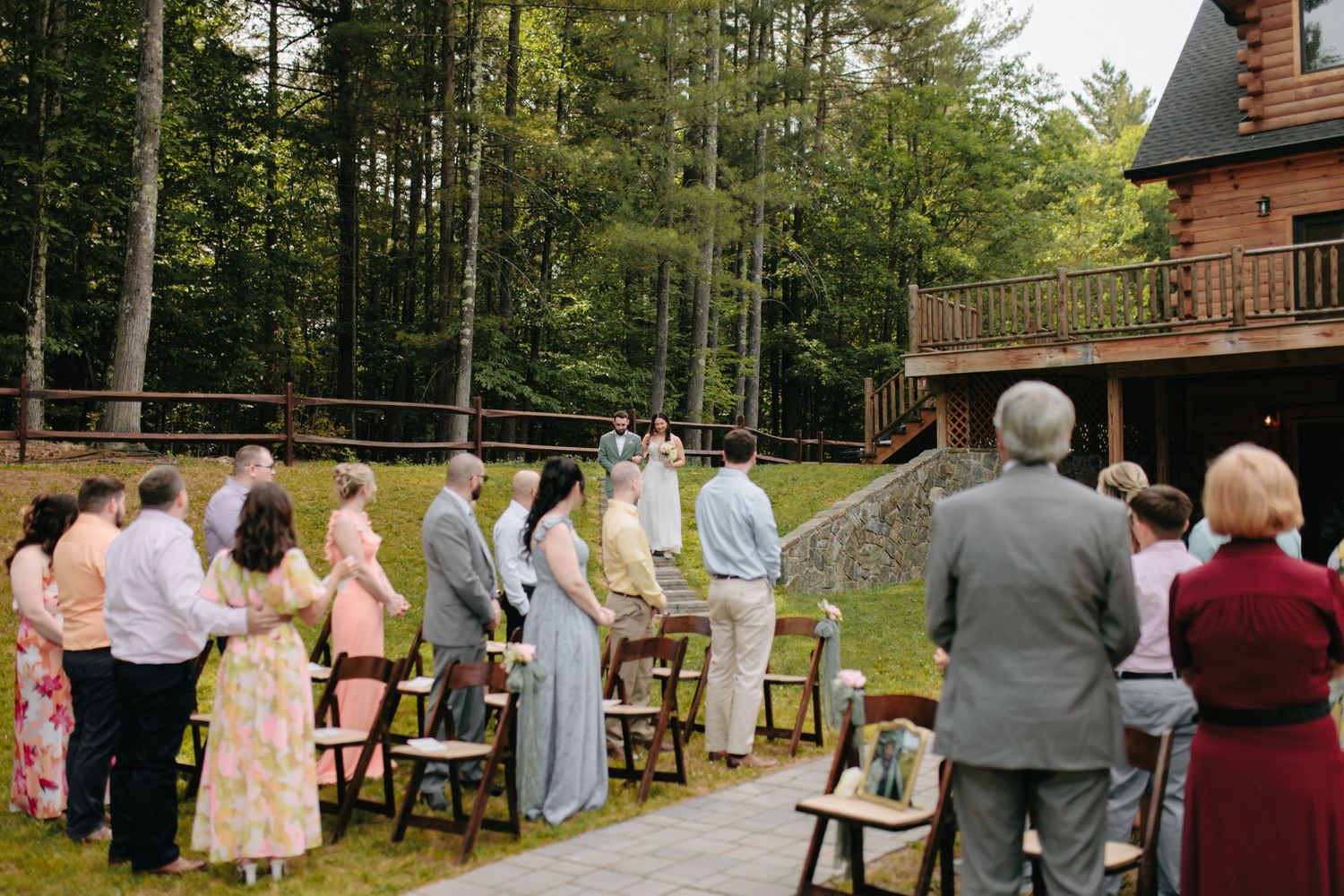 Wedding ceremony guests watch a couple exchange vows in front of a rustic cabin in the woods.