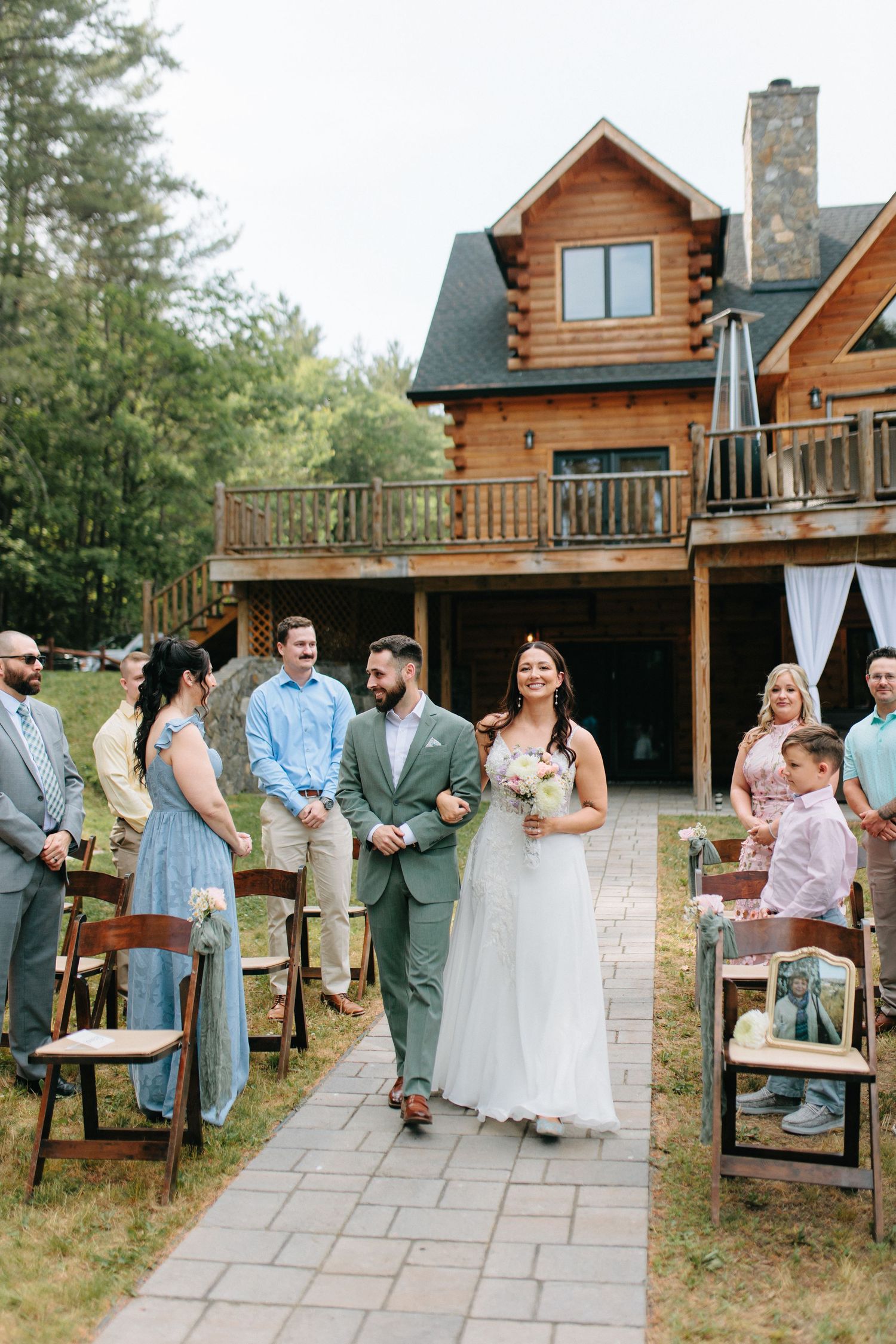 Wedding ceremony setup outside a rustic log cabin with wooden chairs and stone pathway.