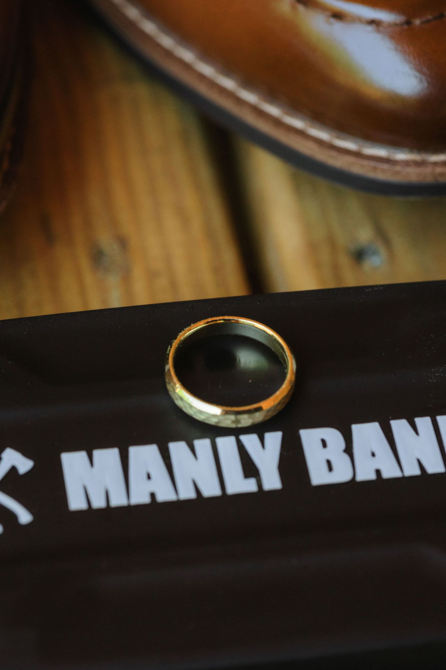 Gold wedding ring displayed on black Manly Bands branded surface with leather shoe in background.