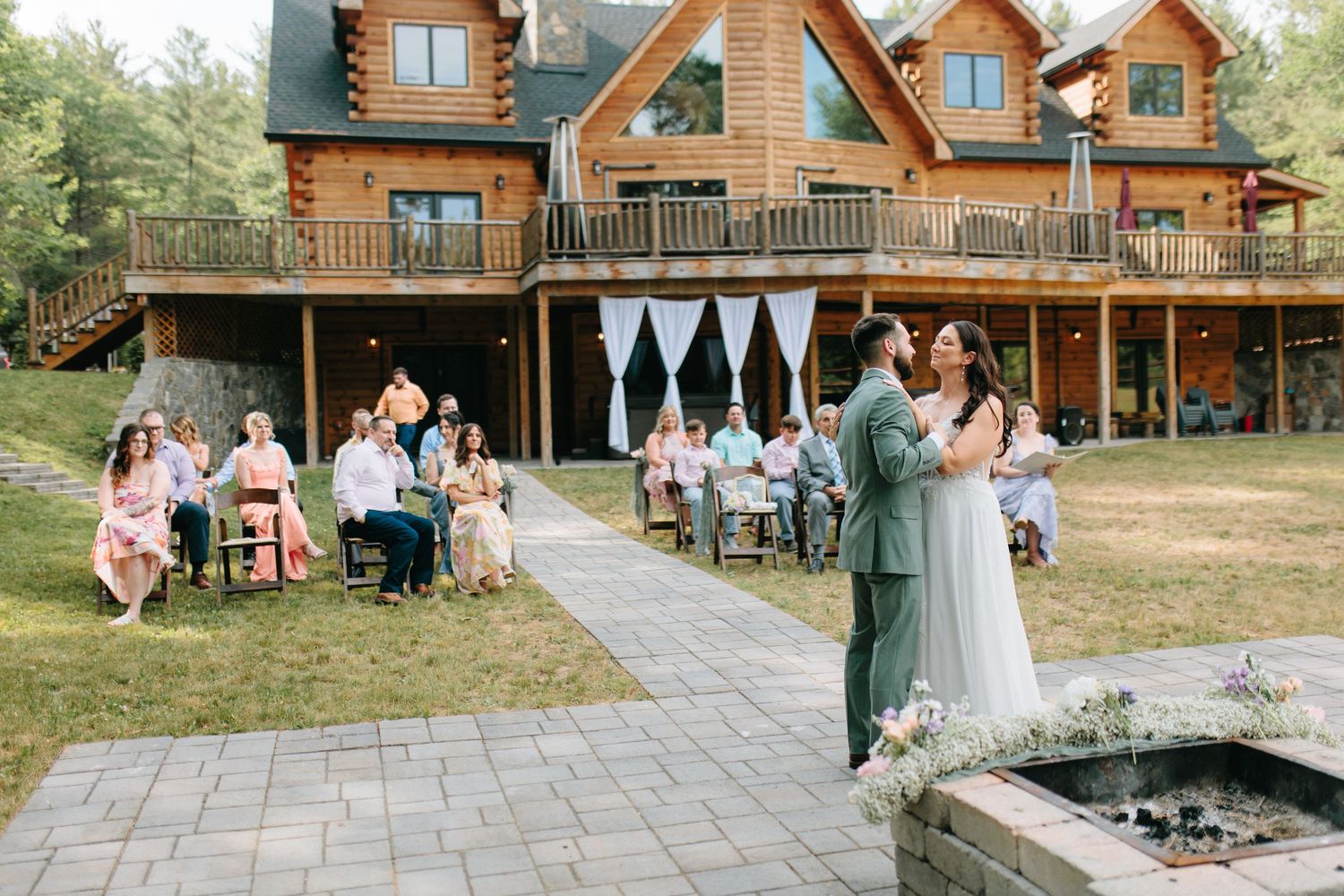 Wedding celebration setup outside a log cabin with stone walkway and wooden seating arrangements.