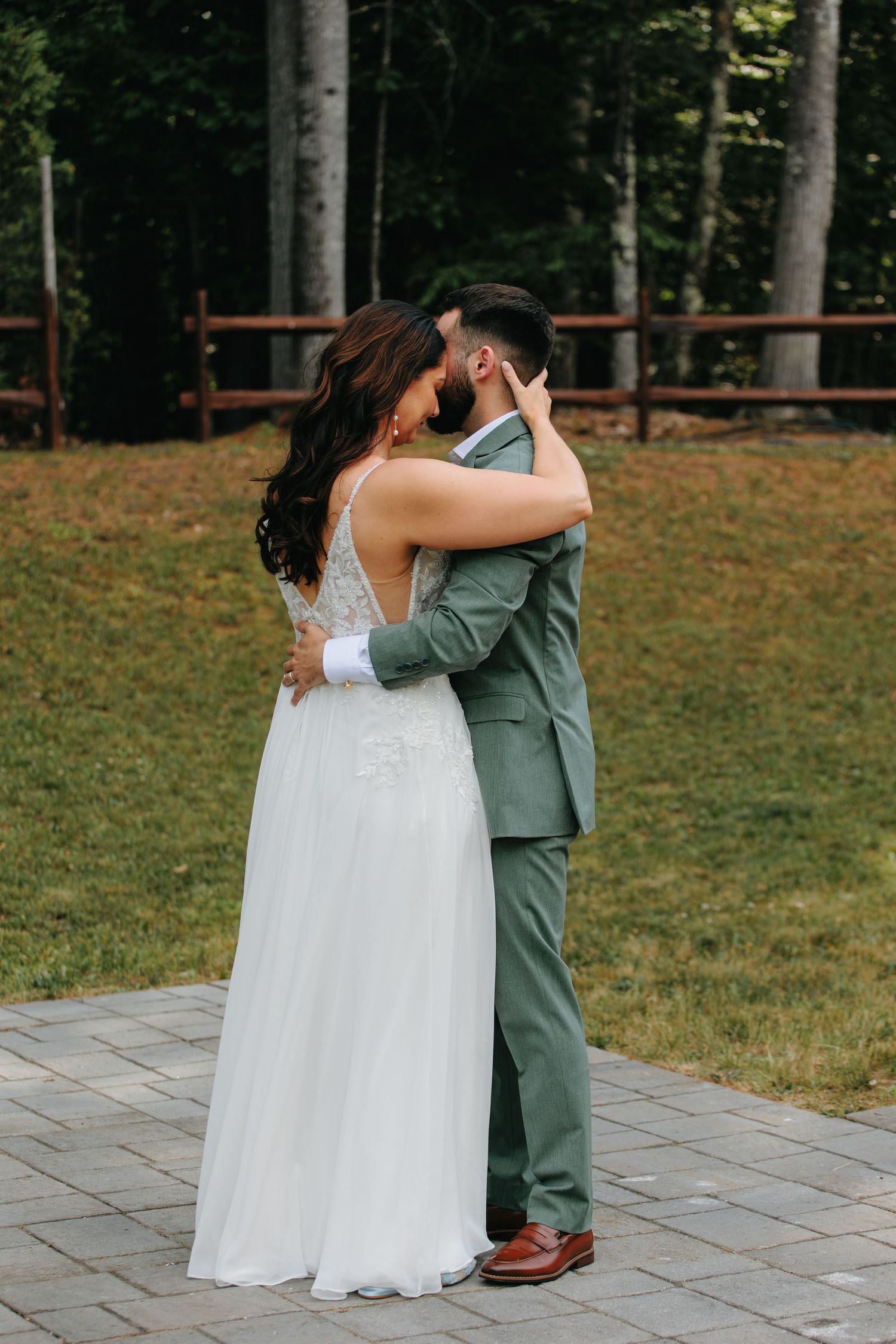 A bride in a white lace gown and groom in sage green suit embrace during their outdoor wedding ceremony.