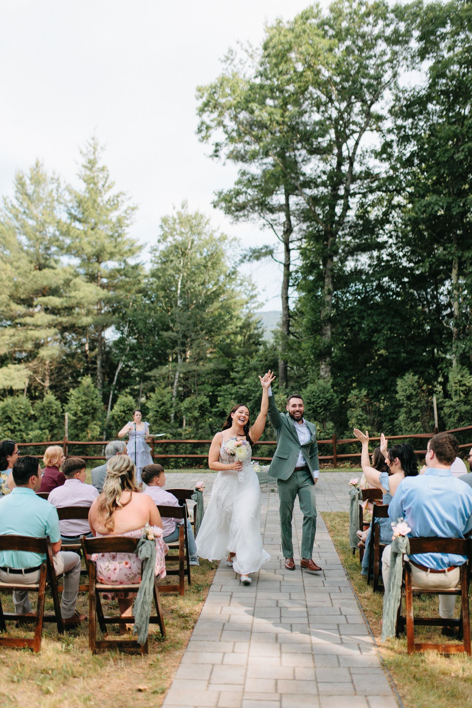 A joyful couple celebrates at an outdoor wedding ceremony on a stone pathway surrounded by tall trees and guests.