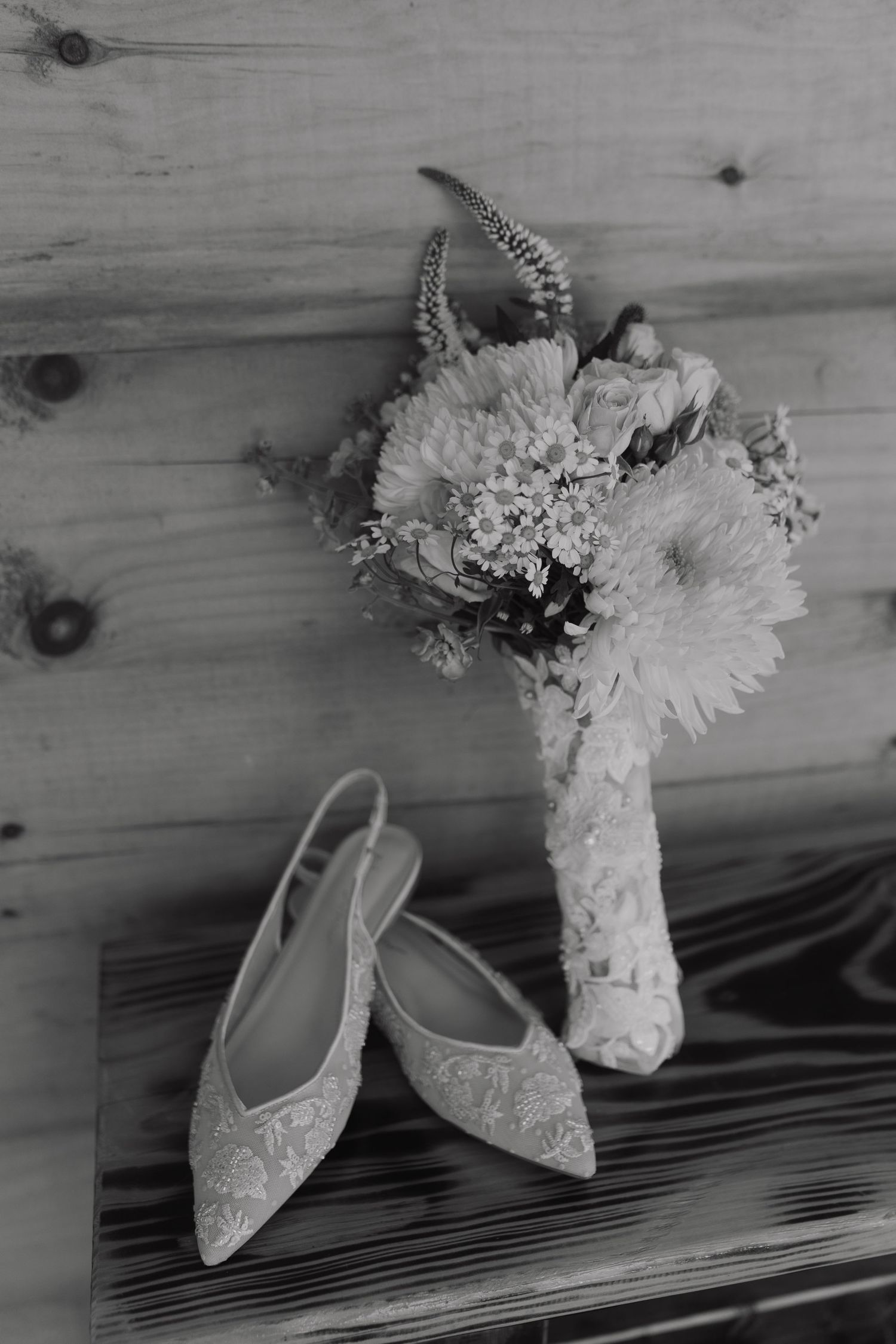 Black and white photo of lace wedding shoes next to a bridal bouquet on wooden surface.