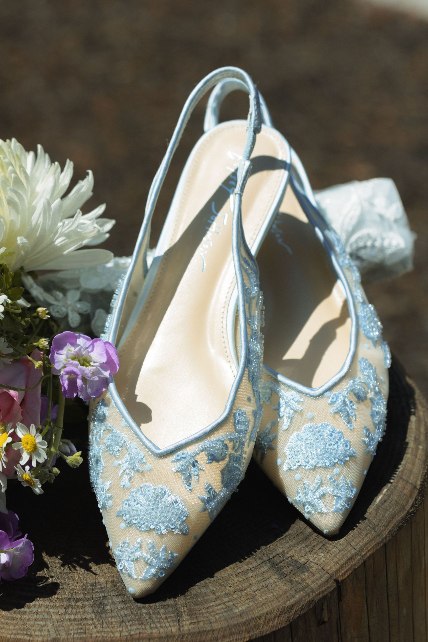 Elegant light blue wedding shoes with lace details displayed with purple and white flowers.