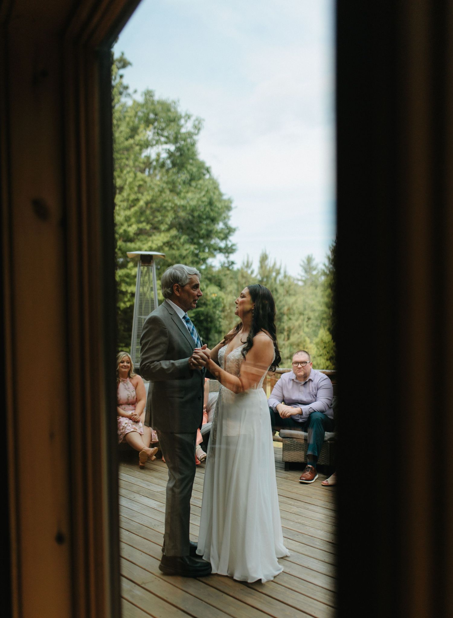 Couple sharing their first dance as newlyweds on a wooden deck surrounded by trees and wedding guests.