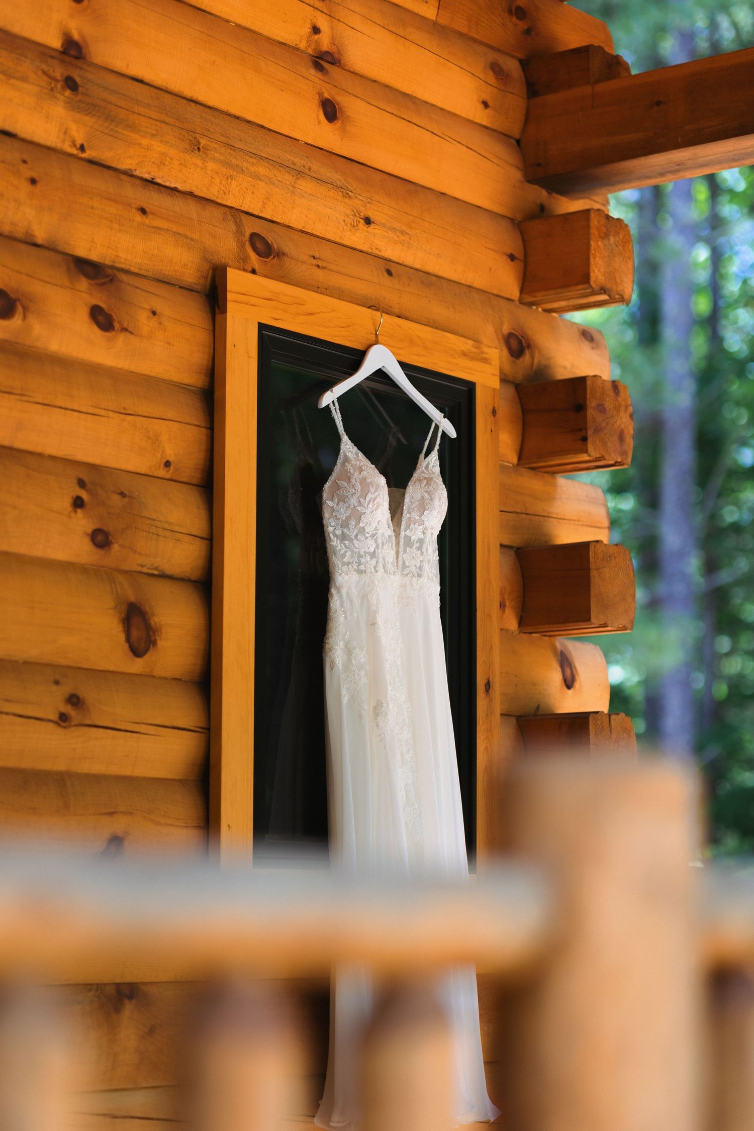 A white lace wedding dress hanging on a rustic log cabin wall.