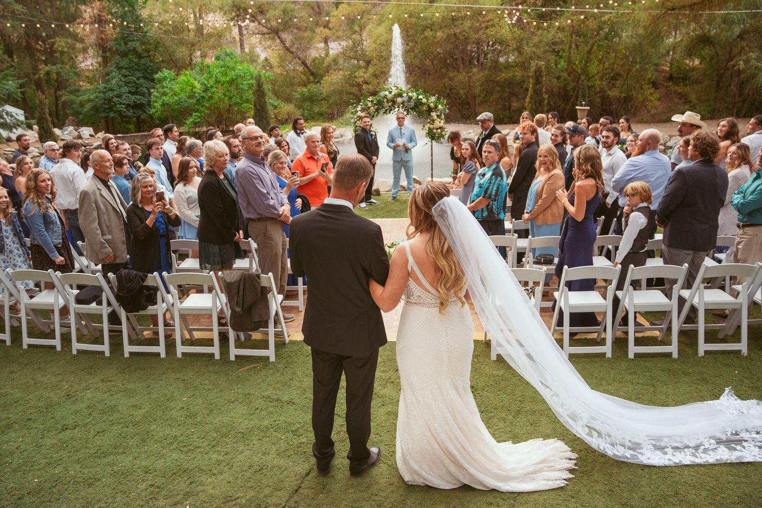 A couple stands in a sunlit meadow wearing wedding attire surrounded by evergreen trees during golden hour.