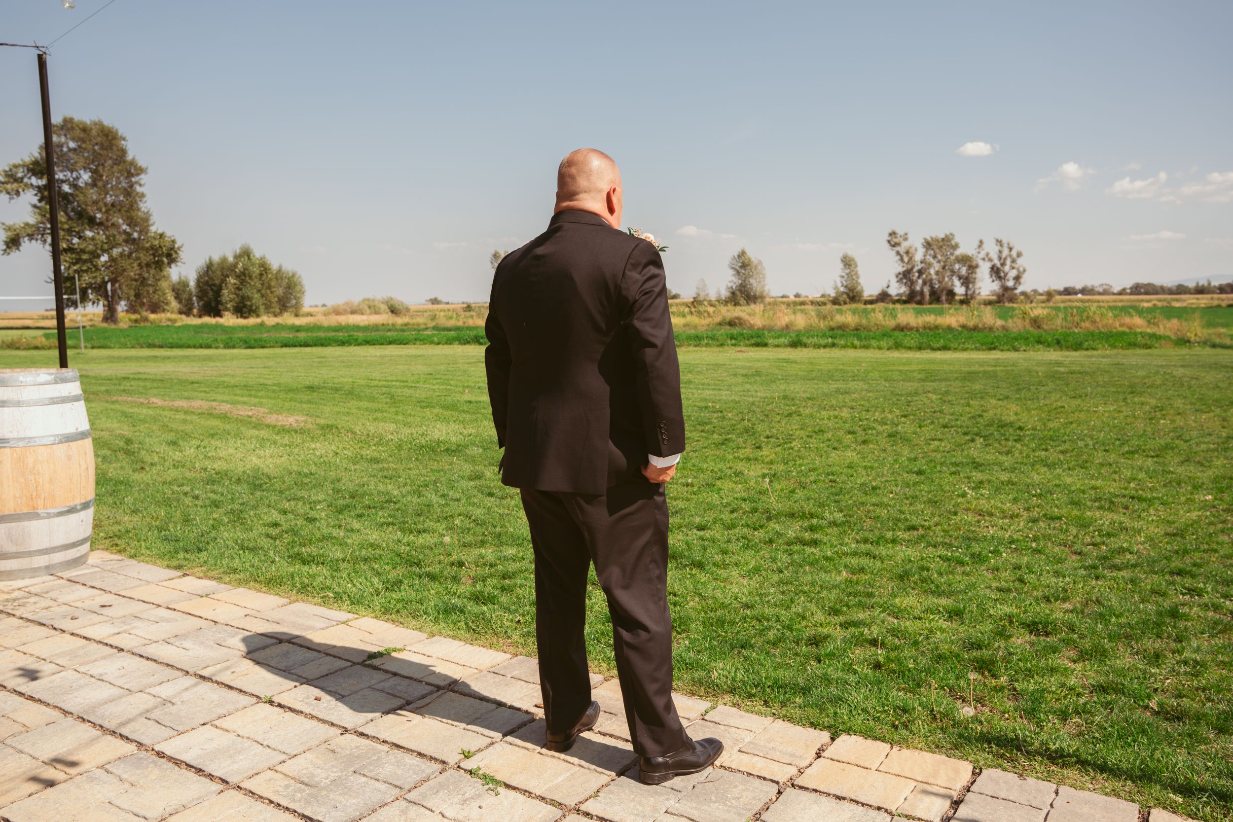 A person in a dark suit stands on stone pavers overlooking a lush green field on a sunny day.