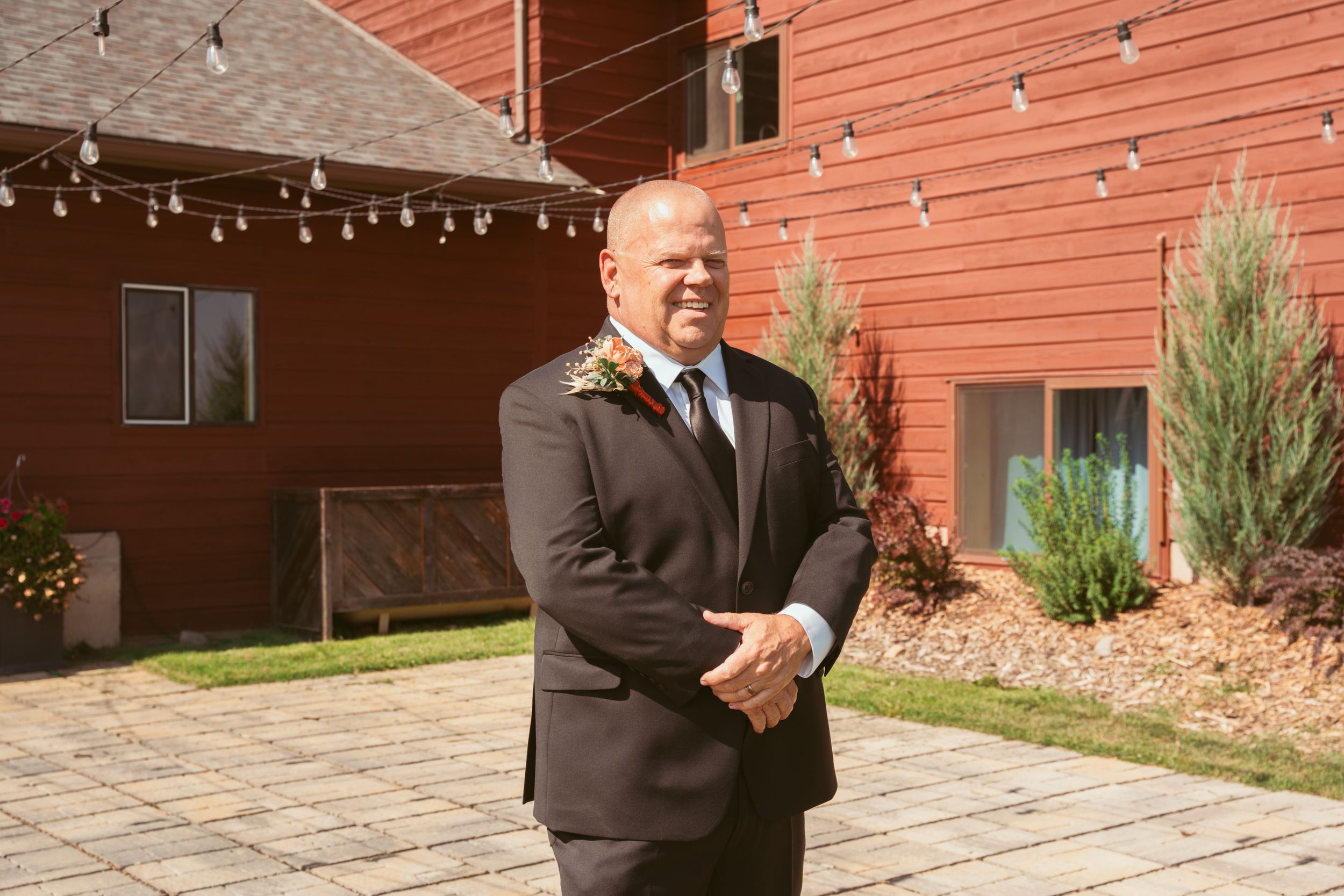 A joyful figure in a formal dark suit stands outside a rustic red barn decorated with string lights.