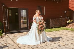 A bride in a stunning white lace dress holds a vibrant autumn-colored bouquet while standing on a patio near a rustic barn.