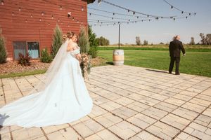 A wedding scene on a paved patio outside a red barn with string lights and a rustic wine barrel decoration.