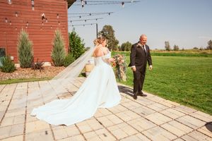 Elegant white wedding dress with long train flows across stone patio at rustic red barn wedding venue.