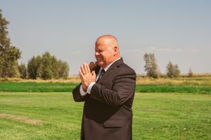 An officiant in a dark suit stands outdoors on green grass gesturing with raised hands during a ceremony.