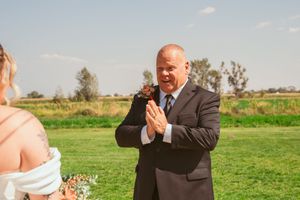 A person in a black suit speaks emotionally while officiating an outdoor ceremony on a grassy field.