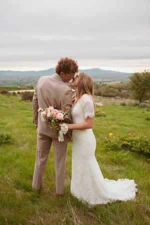 A romantic wedding couple embraces in a green meadow landscape with mountains in the background.